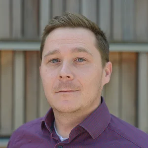 Portrait zeigt Julien Romburg vor einer Holzwand un einem lilafarbenden Hemd.