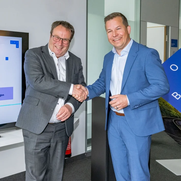 Zwei Männer in Anzug schütteln sich vor einem Bildschirm die Hand, der eine Karte mit Mobilitätsinformationen zeigt.