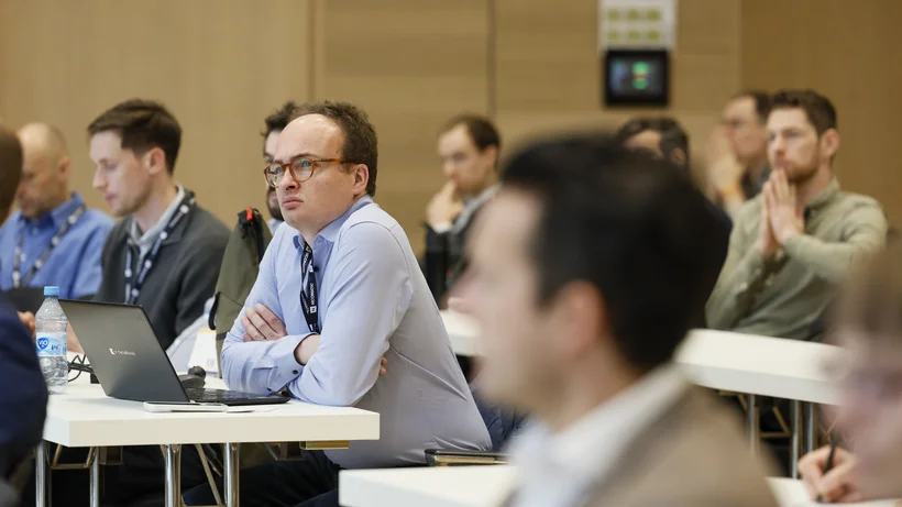 Mehrere Personen sitzen in einem Konferenzraum an Tischen, einige mit Laptops, und hören aufmerksam zu