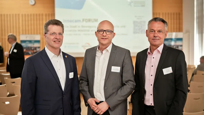 Drei Männer in Anzügen mit Namensschildern stehen in einem Konferenzraum mit Stuhlreihen und einer Präsentationsleinwand im Hintergrund