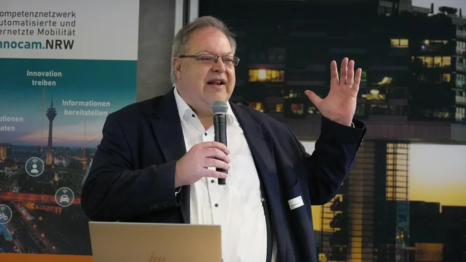 Mann in dunklem Sakko und weißem Hemd hält Mikrofon und gestikuliert mit erhobener Hand vor einem Banner mit dem Schriftzug 'innocam.NRW' und einem Laptop auf dem Pult.