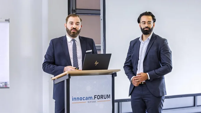 Zwei Männer in Anzügen stehen bei einem Rednerpult mit Laptop, auf dem Schild steht 'innocam.FORUM 18.09.2025'.