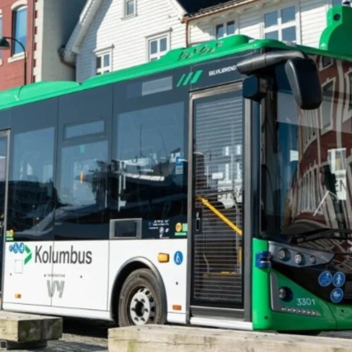 Ein kleiner autonom fahrender Linienbus steht an einer Haltestelle vor städtischer Kulisse.