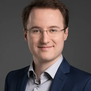Portrait zeigt Christian Frowein, lächelnd mit Brille in einem hellen Hemd und dunklem Jacket vor dunklem Hintergrund.