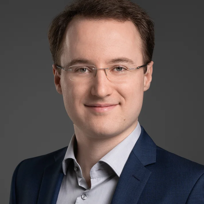 Portrait zeigt Christian Frowein, lächelnd mit Brille in einem hellen Hemd und dunklem Jacket vor dunklem Hintergrund.