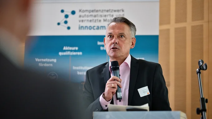 Mann in Anzug mit Mikrofon vor Banner mit Text und Logo, spricht an Rednerpult