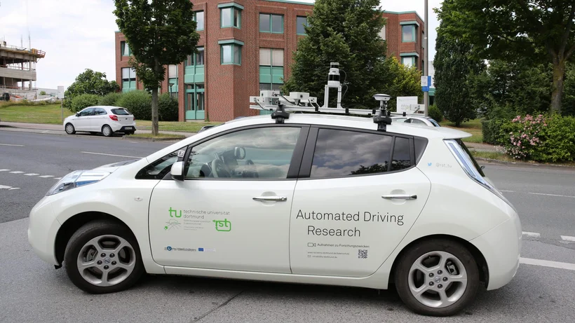 Ein weißes Auto seitlich fotografiert mit Sensoren auf dem Dach und dem Logo der TU Dortmund sowie Automated Driving Research als Aufschrift an den Seitentüren.