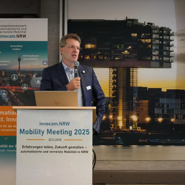 PRof. Dr. Eckstein hält Mikrofon und steht hinter Rednerpult mit Schild 'innocam.NRW Mobility Meeting 2025'. Im Hintergrund Banner mit Text und Grafiken zur automatisierten, vernetzten Mobilität.