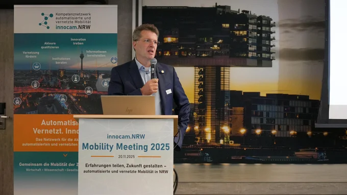 PRof. Dr. Eckstein hält Mikrofon und steht hinter Rednerpult mit Schild 'innocam.NRW Mobility Meeting 2025'. Im Hintergrund Banner mit Text und Grafiken zur automatisierten, vernetzten Mobilität.