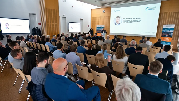 Vortragsraum mit Publikum, das einem Redner an einem Podium und einer Präsentation auf einer Leinwand zuhört