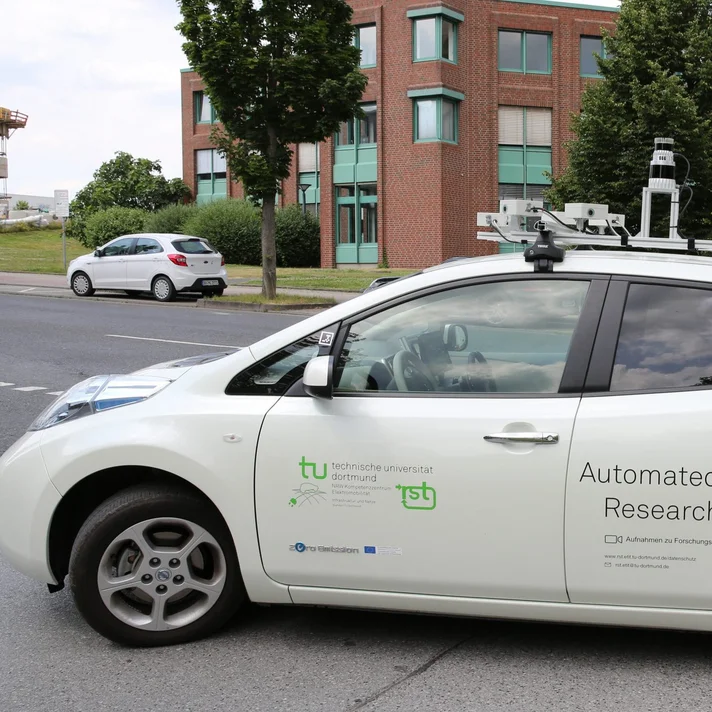 Ein weißes Auto seitlich fotografiert mit Sensoren auf dem Dach und dem Logo der TU Dortmund sowie Automated Driving Research als Aufschrift an den Seitentüren.