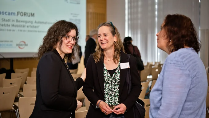 Drei Frauen im Gespräch in einem Veranstaltungsraum mit Stuhlreihen und einer Präsentationsleinwand im Hintergrund