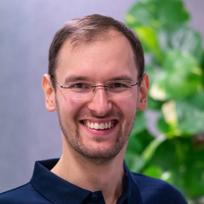 Porträt von Sven Jackisch mit Brille, lächelnd in einem blauen Poloshirt vor einer unscharfen Pflanze im Hintergrund.