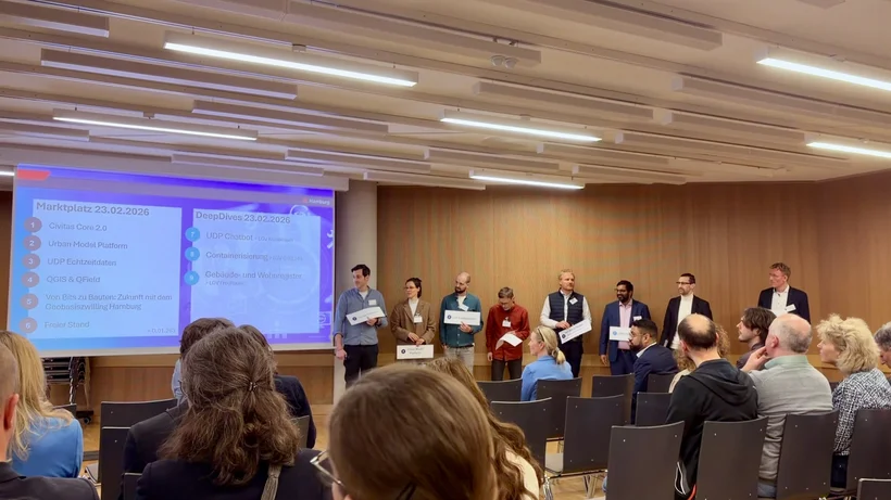 Mehrere Personen stehen in einer Reihe vor einem Publikum in einem Konferenzraum mit Holzverkleidung und einer Präsentation an der Wand.