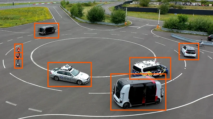 Mehrere autonome Fahrzeuge und zwei Personen in Warnwesten auf einem Testgelände. Die Fahrzeuge, darunter Autos und ein Shuttlebus, sind mit Sensoren ausgestattet und fahren auf einer markierten Kreisbahn. Alle Objekte sind mit orangefarbenen Rechtecken markiert.