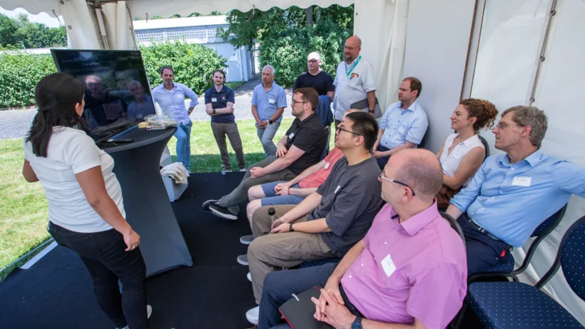 Gruppe von Menschen sitzt und steht in einem Zelt, eine Frau präsentiert vor einem Bildschirm
