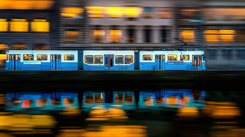 Hellblaue-weiße Straßenbahn mit mehreren Fenstern fährt vor einem Gebäude mit gelb beleuchteten Fenstern, Spiegelung im Wasser sichtbar.