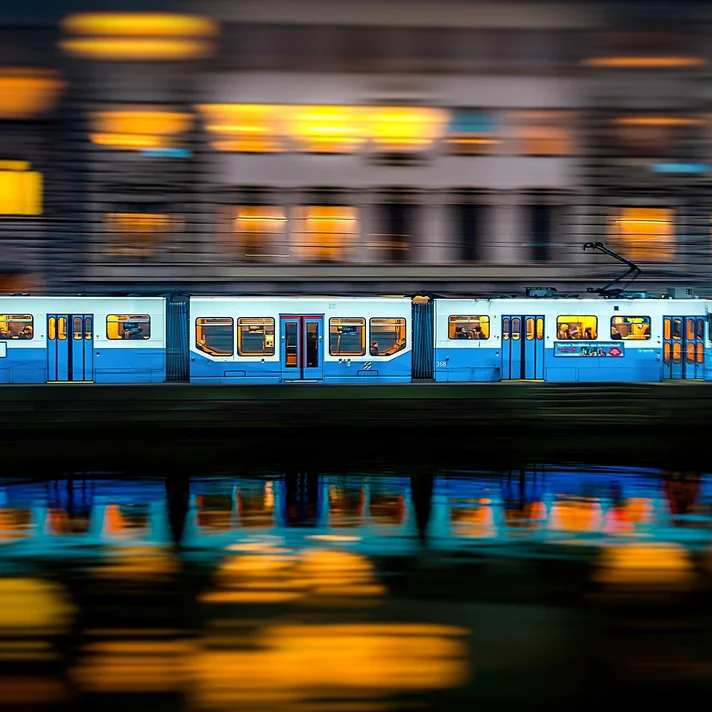 Hellblaue-weiße Straßenbahn mit mehreren Fenstern fährt vor einem Gebäude mit gelb beleuchteten Fenstern, Spiegelung im Wasser sichtbar.
