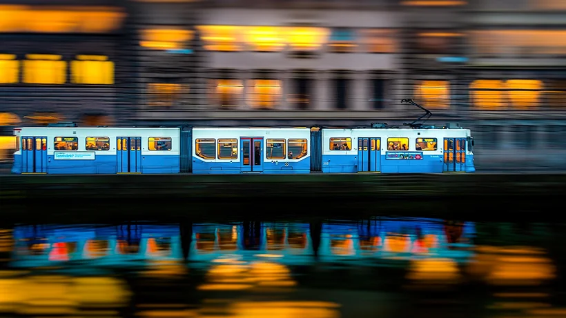 Hellblaue-weiße Straßenbahn mit mehreren Fenstern fährt vor einem Gebäude mit gelb beleuchteten Fenstern, Spiegelung im Wasser sichtbar.