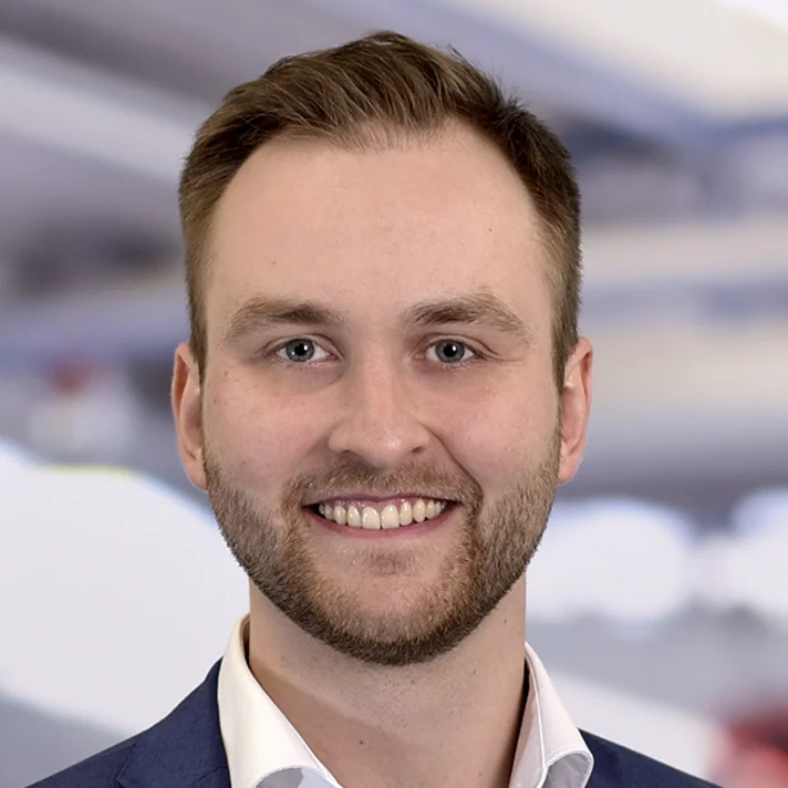 Porträt von Matthis Hötter, Innovationsmanager, mit kurzem Haar, Bart und blauem Sakko, freundlich lächelnd vor hellem, unscharfem Hintergrund.