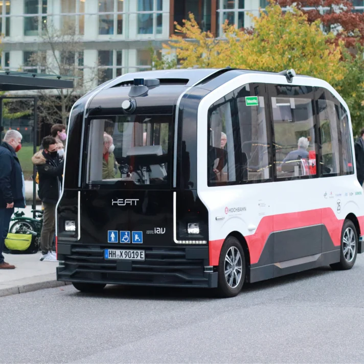 Autonom fahrender Shuttlebus an einer Haltestelle in urbaner Umgebung, Passanten stehen am Straßenrand.