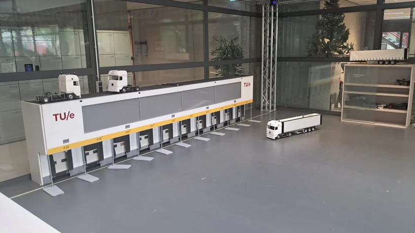 Modell einer automatisierten Lager- oder Produktionsanlage mit mehreren weißen Containern und einem Lkw im Innenraum eines modernen Gebäudes.