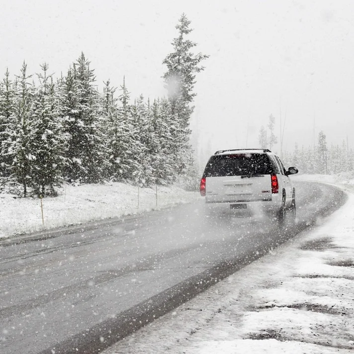 Man sieht ein weißes Auto von hinten, das auf einer verschneiten Straße fährt, links sieht man Nadelbäume und e