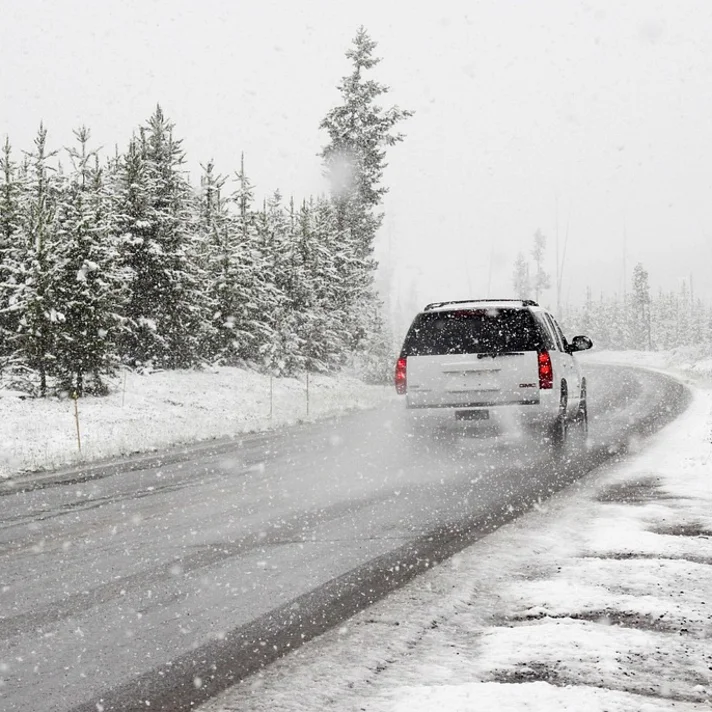 Man sieht ein weißes Auto von hinten, das auf einer verschneiten Straße fährt, links sieht man Nadelbäume und e