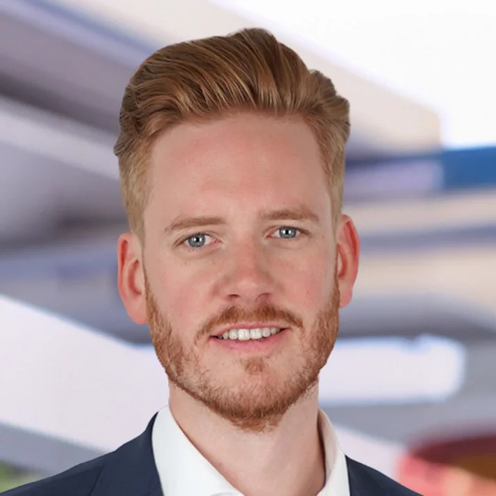 Portrait David Haberle „Porträtfoto lächelnd mit kurzem, nach hinten gestyltem rötlich-blondem Haar und gepflegtem Bart. Er trägt ein weißes Hemd und ein dunkles Sakko. Der Hintergrund ist unscharf und zeigt helle, moderne Architektur.“