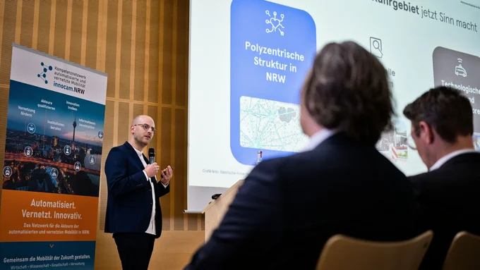 Mann mit Mikrofon hält Vortrag vor Präsentationsfolie und Banner zu vernetzter Mobilität im Ruhrgebiet, zwei Männer im Vordergrund sitzen auf Stühlen