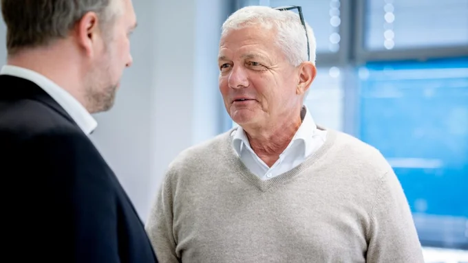 Zwei Männer im Gespräch, einer trägt einen hellen Pullover und ein weißes Hemd, der andere einen dunklen Anzug mit weißem Hemd.