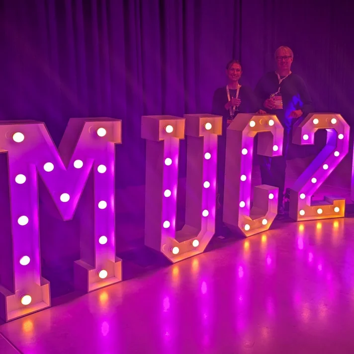 In pinkes Licht getauchte Szene mit  von einzelnen Glühlampen von innen heraus beleuchteten Buchstaben die MUC2025 buchstabieren und etwa seinen Meter hoch sind, dahinter posieren zwei Personen.