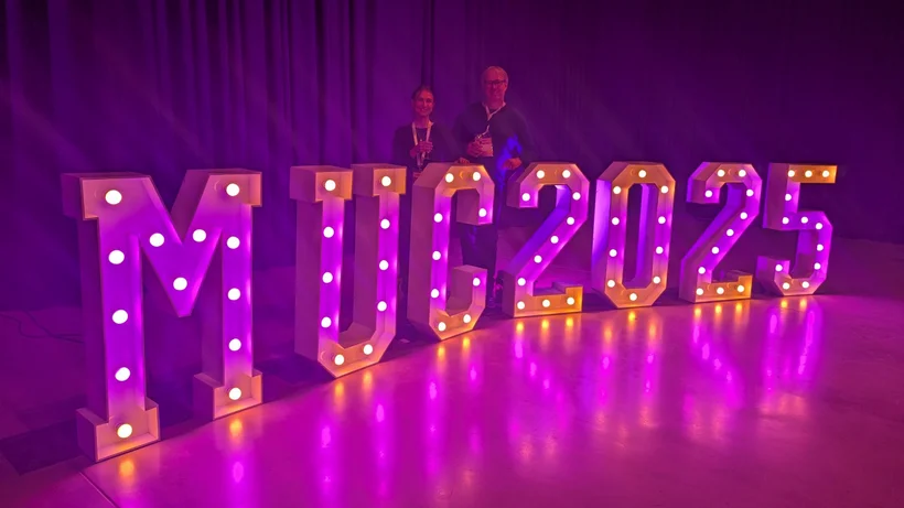 In pinkes Licht getauchte Szene mit  von einzelnen Glühlampen von innen heraus beleuchteten Buchstaben die MUC2025 buchstabieren und etwa seinen Meter hoch sind, dahinter posieren zwei Personen.