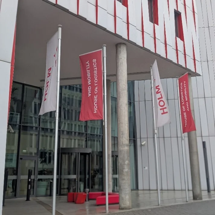 Fahnen mit der Aufschrift 'HOLM' und 'HOUSE OF LOGISTICS AND MOBILITY' vor einem modernen Gebäude mit vertikalen roten und weißen Fassadenelementen.