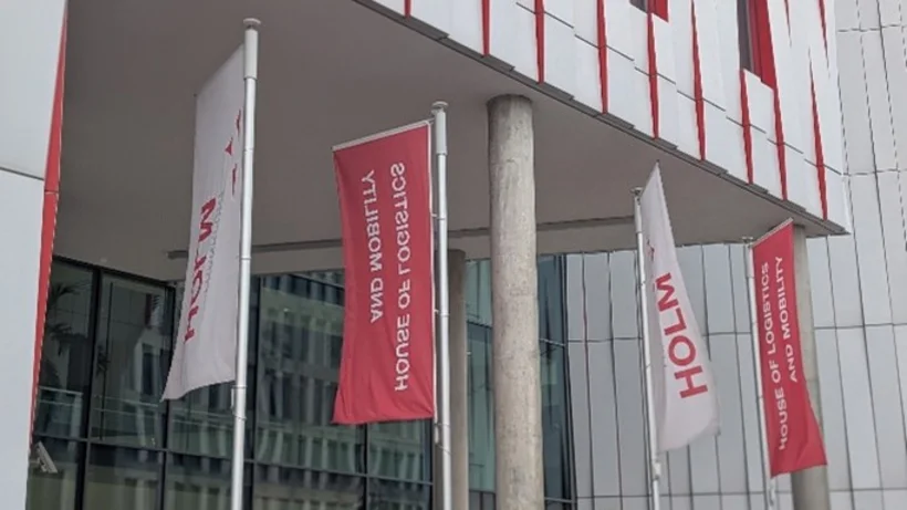 Fahnen mit der Aufschrift 'HOLM' und 'HOUSE OF LOGISTICS AND MOBILITY' vor einem modernen Gebäude mit vertikalen roten und weißen Fassadenelementen.