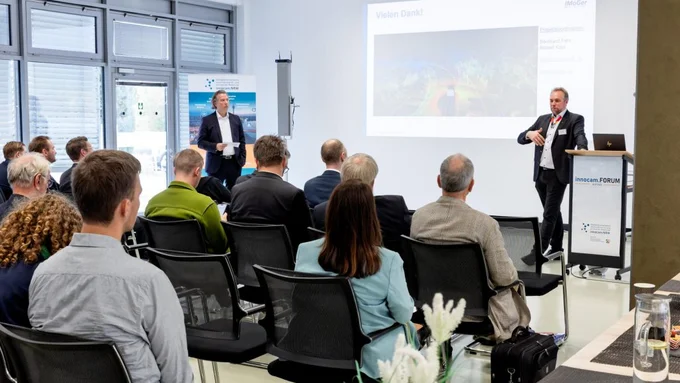 Zwei Männer halten eine Präsentation vor sitzendem Publikum in einem hellen Raum mit Fensterfront und Projektionsleinwand.