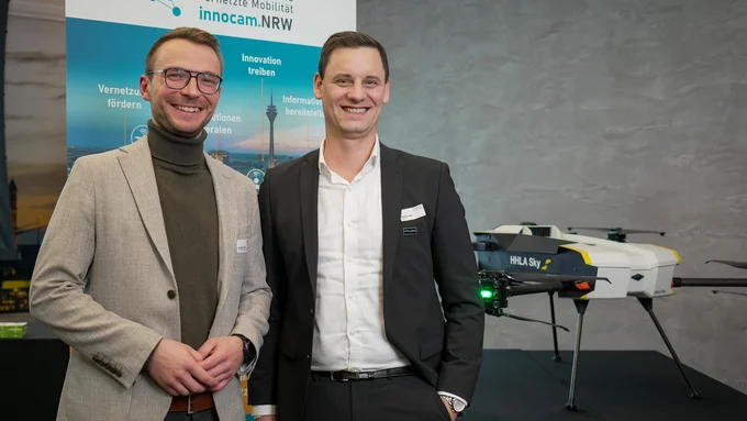 Zwei Männer in Businesskleidung stehen vor einem Banner mit dem Logo von innocam.NRW und einem weißen Drohnenmodell mit der Aufschrift 'HHLA Sky'.
