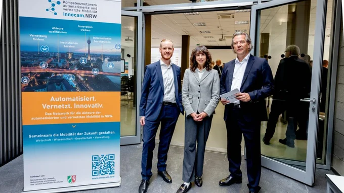 Drei Personen in Businesskleidung stehen nebeneinander vor einem geöffneten Glastürrahmen neben einem Roll-up mit dem Titel 'innocam.NRW' und dem Slogan 'Automatisiert. Vernetzt. Innovativ.'.
