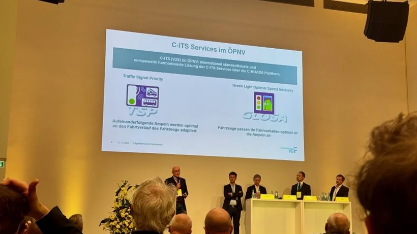 Auf einer beleuchteten Bühne steht ein Moderator und recht dabene sitzen hinter einer weißen theke 4 Männer in dunklen Anzügen aus einem Panel zum Thema C-ITS Services, eine Folie dazu ist auf der Rückwand des Raumes projiziert, im Vordergrund sind Zuschauer von hinten in Sitzreihen im Bild.