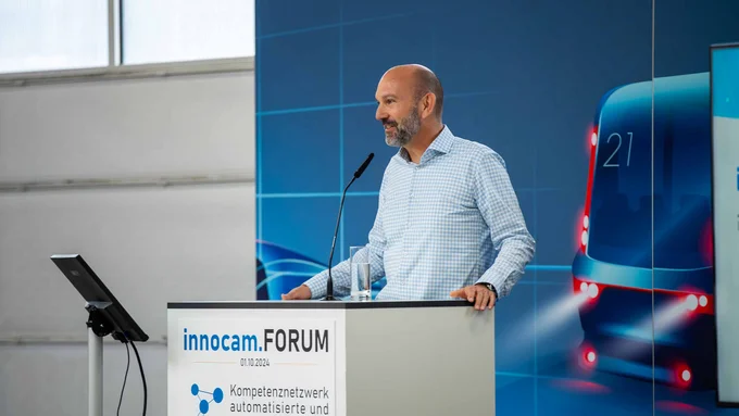 Ein Mann im karierten Hemd spricht am Pult mit dem Logo „innocam.FORUM“.