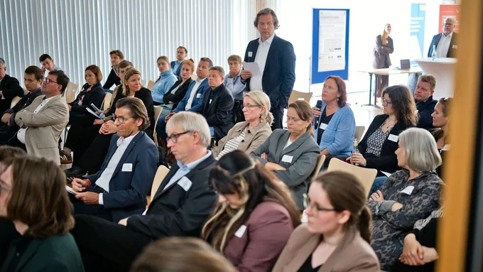 Gruppe von Menschen in einem hellen Raum bei einer Veranstaltung, eine Person steht mit Mikrofon und spricht