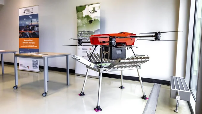 Roter Drohnenprototyp mit vier Rotoren und Metallgestell steht in einem hellen Raum neben zwei Rolltischen und Bannern.