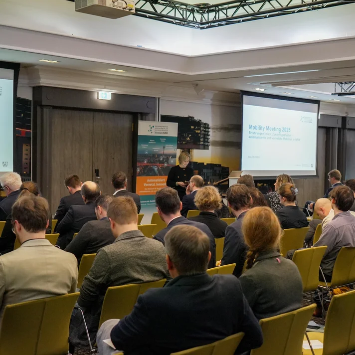 Teilnehmende sitzen in einem Konferenzraum bei der innocam.NRW Mobility Meeting Veranstaltung, Blick auf die Rückseite der Personen und zwei Projektionsflächen vorne.