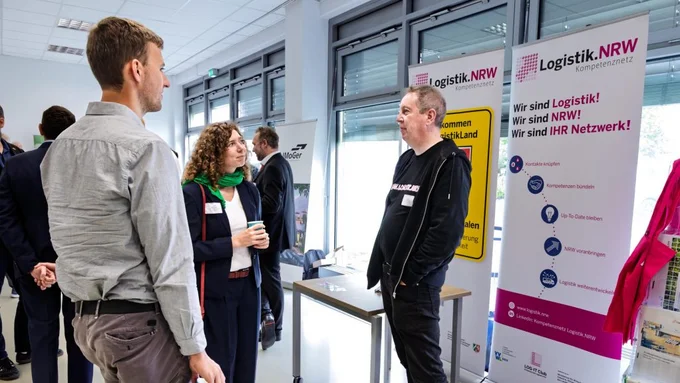 Mehrere Personen stehen in einem hellen Raum vor zwei aufrechten Bannern mit den Überschriften 'Logistik.NRW' und 'Logistik.NRW Kompetenznetz'.