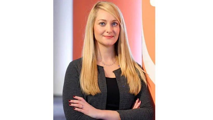 Porträtfoto von Jana Kalbfleisch, einer lächelnden jungen Frau mit langen blonden Haaren und verschränkten Armen, Business Kleidung tragend, der Hintergrund ist unscharf und im Orangeton gehalten.