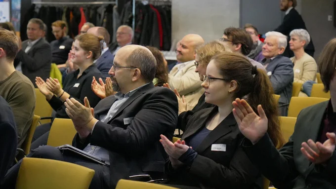 Mehrere Personen sitzen in einem Konferenzraum und klatschen, im Hintergrund Garderobe mit Jacken zu sehen.