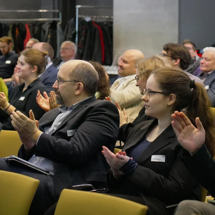 Mehrere Personen sitzen in einem Konferenzraum und klatschen, im Hintergrund Garderobe mit Jacken zu sehen.