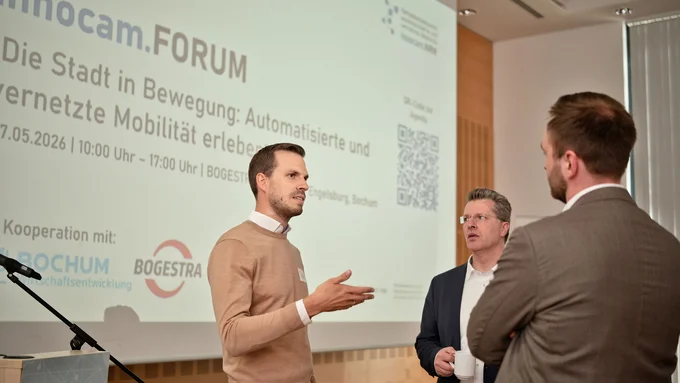 Drei Männer im Gespräch vor einer Projektionswand mit Text zum inncam.FORUM und Logos von Bochum und BOGESTRA