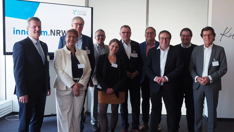 Zehn Personen stehen in Businesskleidung vor einem Bildschirm mit dem Logo von innocam.NRW und posieren für ein Gruppenfoto.  Caption: Mitglieder des Mobilitätsrats von innocam.NRW bei einem offiziellen Treffen in Köln.