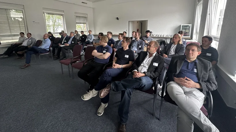 Eine Gruppe von etwa zwanzig Personen sitzt in einem Konferenzraum und hört einer Präsentation zu. Die Teilnehmer tragen Namensschilder und sind in Reihen auf Stühlen angeordnet. Der Raum ist hell mit großen Fenstern, durch die Tageslicht einfällt.